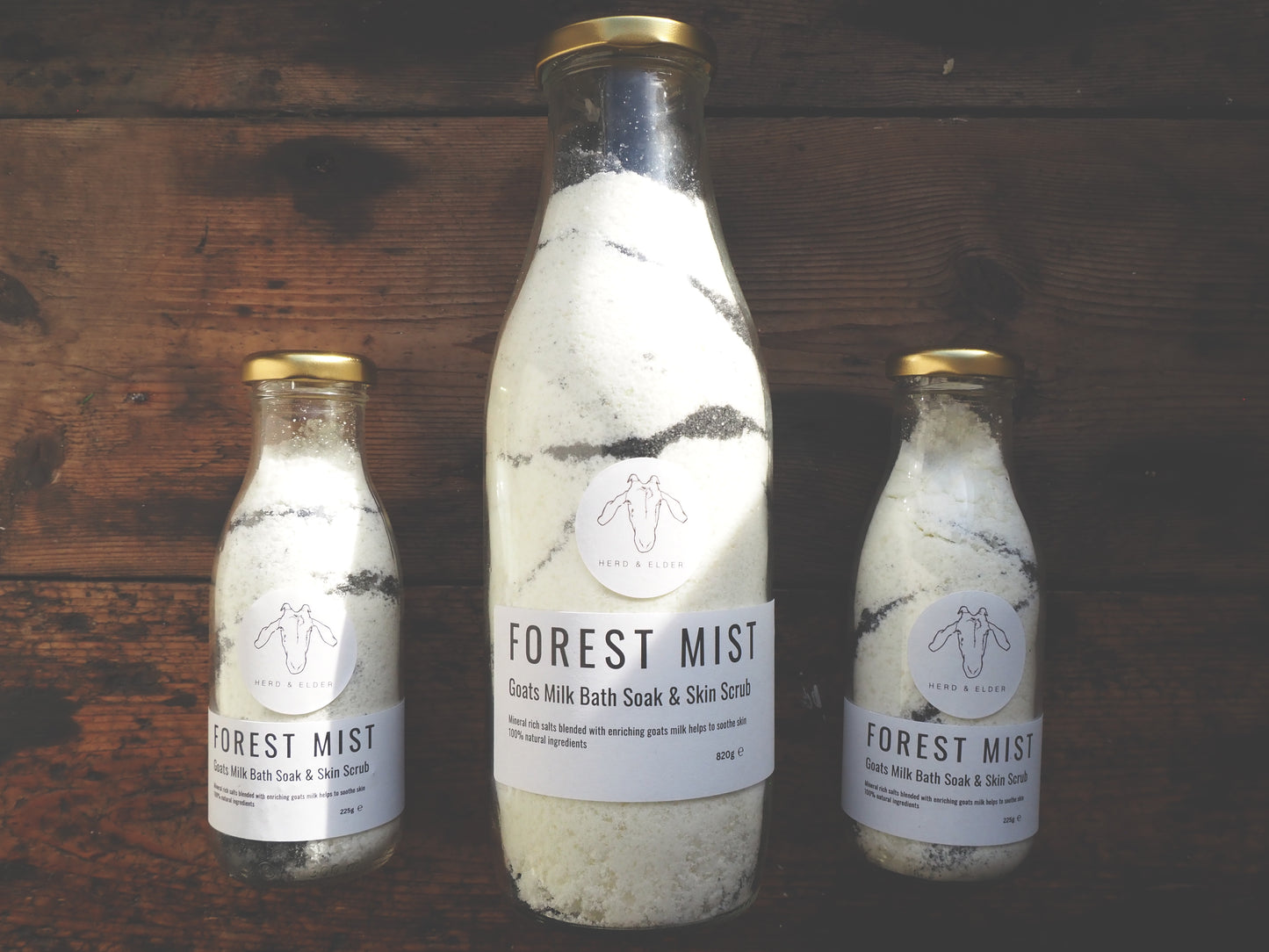 Goat Milk Bath Salts in clear glass bottles labeled 'Forest Mist' on wooden background, showcasing the skin scrub.