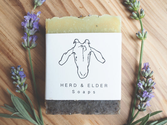 Goats milk soap wrapped in rustic label with goat illustration and surrounded by lavender flowers on a wooden surface.